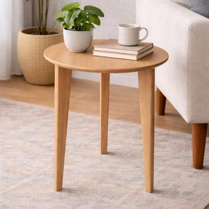May include: A small, round wooden side table with three angled legs. A small potted plant, two books, and a mug sit on top. The table is on a patterned rug, with a sofa and a large potted plant in the background.