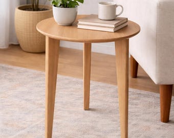 Op de afbeelding: Een kleine, ronde houten bijzettafel met drie schuine poten. Een kleine pot met plant, twee boeken en een mok staan erop. De tafel staat op een patroonkleed, met een bank en een grote potplant op de achtergrond.