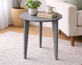 Op de afbeelding: Een grijze ronde bijzettafel met drie schuine poten. Een kleine plant in een pot, boeken en een mok staan erop. Een grotere plant in een gevlochten mand en een bank met groene kussens staan op de achtergrond.