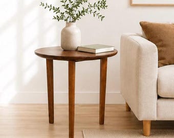 Op de afbeelding: Een ronde, donkerbruine houten bijzettafel met drie taps toelopende poten. Een beige vaas met groene takken en een boek staan op de tafel. De tafel staat naast een beige bank met een bruin kussen.