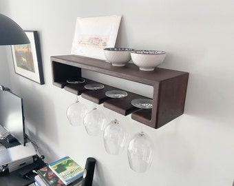 Op de afbeelding: Een aan de muur gemonteerd wijnrek van donkerbruin hout, met vier wijnglazen. Twee witte kommen met blauwe patronen staan op de bovenste plank. Het rek heeft een modern, minimalistisch ontwerp.