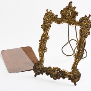 Puede incluir: Marco de fotos dorado de estilo antiguo con intrincados adornos y un soporte decorativo. Un papel marrón y beige está al lado del marco. El marco está vacío, listo para una foto o una obra de arte.