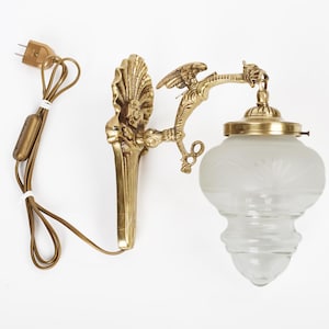 Puede incluir: Aplique de pared de latón dorado con una pantalla de cristal esmerilado. El aplique presenta elementos escultóricos detallados, incluyendo una figura alada y un diseño en forma de concha. El cable eléctrico y el enchufe son visibles.