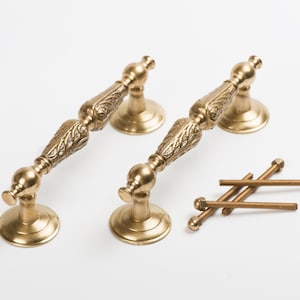 Può includere: Due maniglie per ante di armadio dorate con disegni decorativi e un set di quattro viti.