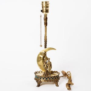 May include: A vintage brass table lamp featuring a crescent moon design with a couple figurine. The lamp has a decorative base with teal accents and ornate detailing. The lamp is approximately 46 cm tall.