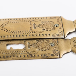 May include: Two antique brass door plates with an ornate design. The plates feature a floral pattern, a vase, and a profile of a person. The plates have a keyhole cutout.