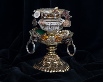 May include: A gold metal jewelry stand with a round top and three legs. The stand is decorated with a white ceramic bowl and a variety of earrings, including pearl, silver, and gold.