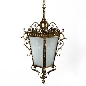 May include: A brass pendant light fixture with a frosted glass shade and intricate scrollwork details. The fixture has a square shape and hangs from a chain.