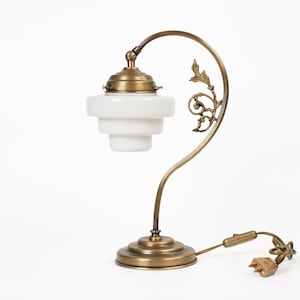 Puede incluir: Lámpara de escritorio antigua de latón con una pantalla de cristal blanco escalonada. La lámpara presenta un brazo curvo con detalles decorativos de hojas y una base redonda. La lámpara tiene un cable y un enchufe para la alimentación.