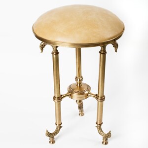 Puede incluir: Pequeña mesa auxiliar de estilo antiguo con una tapa redonda acolchada de color beige claro. La mesa tiene una estructura metálica de color latón con tres patas y detalles decorativos. La mesa mide unos 61 cm de alto.