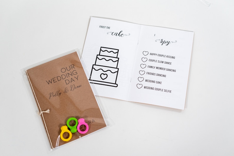 Puede incluir: Un folleto de papel kraft marr&oacute;n con el texto "OUR WEDDING DAY Polly & Dean" en la portada. El folleto est&aacute; envuelto en pl&aacute;stico transparente y atado con una cuerda. Se incluyen tres anillos de pl&aacute;stico de colores. El folleto est&aacute; abierto para revelar una p&aacute;gina con una ilustraci&oacute;n en blanco y negro de un pastel de bodas y el texto "FROST THE cake". La p&aacute;gina opuesta tiene el texto "I Spy" y una lista de art&iacute;culos relacionados con la boda para encontrar.