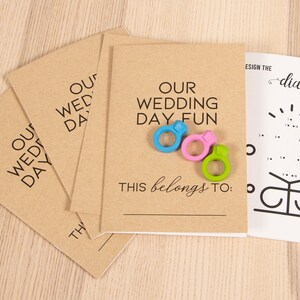 May include: Four brown cardstock envelopes with the text "Our Wedding Day Fun" and "This belongs to:" printed on them. Three colorful plastic rings and a black plastic ring are on the envelopes. A white sheet of paper with a connect-the-dots diamond ring design is in the background.