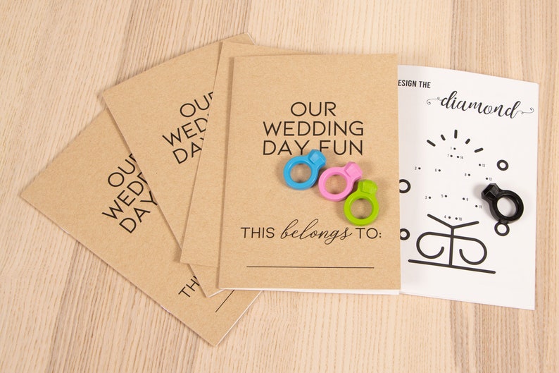May include: Four brown cardstock envelopes with the text "Our Wedding Day Fun" and "This belongs to:" printed on them. Three colorful plastic rings and a black plastic ring are on the envelopes. A white sheet of paper with a connect-the-dots diamond ring design is in the background.