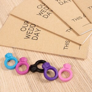 May include: Five colorful ring-shaped objects with a diamond-shaped design on top. The rings are blue, pink, black, purple, and pink. The rings are arranged in a row on a wooden surface.  The rings are next to a stack of brown notebooks with the text "Our Wedding Day" printed on the cover.
