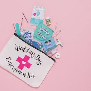 May include: A white canvas Wedding Day Emergency Kit bag with a pink cross and heart graphic, filled with various small items. Includes Wet Ones, a razor, deodorant, bobby pins, and other essentials. The bag is on a pink background.