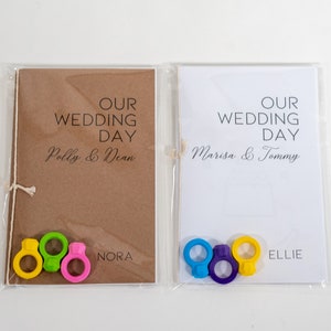 Puede incluir: Dos tarjetas de d&iacute;a de boda con los nombres Polly & Dean y Marisa & Tommy impresos en ellas. Cada tarjeta tiene una cuerda atada alrededor y tres anillos coloridos unidos a la cuerda. Los anillos son amarillos, verdes y rosas en una tarjeta y azules, morados y amarillos en la otra. Las tarjetas tambi&eacute;n tienen los nombres Nora y Ellie impresos en ellas.
