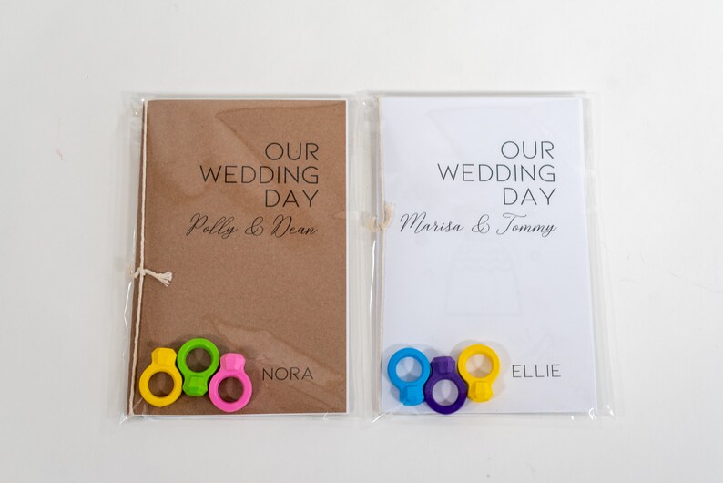 Puede incluir: Dos tarjetas de d&iacute;a de boda con los nombres Polly & Dean y Marisa & Tommy impresos en ellas. Cada tarjeta tiene una cuerda atada alrededor y tres anillos coloridos unidos a la cuerda. Los anillos son amarillos, verdes y rosas en una tarjeta y azules, morados y amarillos en la otra. Las tarjetas tambi&eacute;n tienen los nombres Nora y Ellie impresos en ellas.
