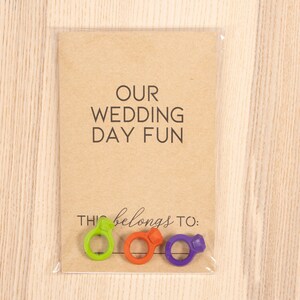 May include: A brown card with the text "OUR WEDDING DAY FUN" and the text "THIS belongs to:" with three colorful ring-shaped objects below the text.