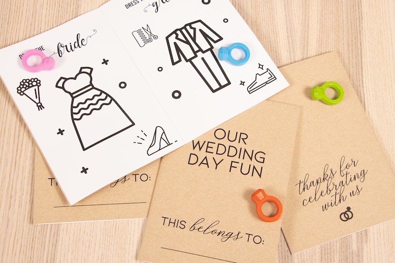 May include: Two brown cardstock paper booklets with the text "OUR WEDDING DAY FUN" and "thanks for celebrating with us". Each booklet has a blank space for a name and a ring-shaped object.
