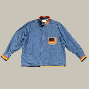 May include: A long-sleeve blue denim button-down shirt featuring a collar with red, orange, and black patterned accents. The shirt includes a chest pocket with a black, orange, and yellow graphic, and matching striped cuffs in red, orange, and black.