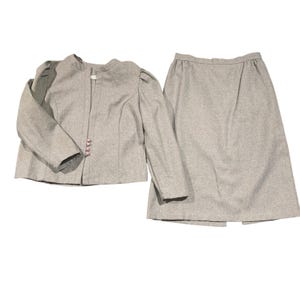 May include: A light gray skirt suit. The jacket has long sleeves, a round collar, and three pink buttons. The skirt is knee-length. The suit is made of a textured fabric.