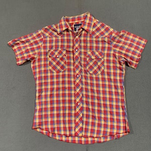 May include: Red, yellow, and blue plaid short-sleeve shirt with two chest pockets and a button-down front. The shirt has a classic Western style with a collared neckline. The brand name "Wrangler" is visible on the label.