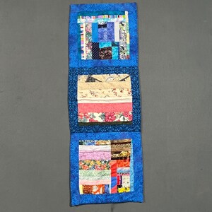 May include: A handmade quilted wall hanging with three rectangular sections. Each section features a patchwork design with various fabric patterns and colors, including blues, pinks, and floral prints. The piece is framed by a blue patterned border.