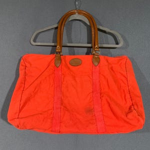 May include: Bright orange duffel bag with brown leather handles and accents. The bag has a rectangular shape with a top zipper closure and two vertical red straps. The bag is hanging on a gray hanger.