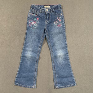 May include: Blue denim jeans featuring a classic five-pocket design with a button and zipper closure. The front of the jeans is decorated with embroidered heart patterns in shades of pink, light blue, and white. A Cherokee brand label is visible.