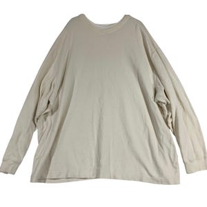 May include: A long-sleeved, cream-colored shirt with a crew neck. The shirt has a relaxed fit and ribbed cuffs at the wrists. The fabric appears to be a soft, knit material. The shirt is laid flat on a white surface.