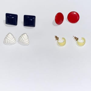 May include: An assortment of earrings in various styles. There are two pairs of navy blue square studs, two pairs of red circular studs, two pairs of white knot-shaped studs, and two pairs of gold-colored, crescent-shaped hoop earrings.