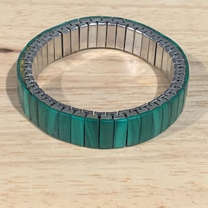 May include: A green malachite watch band with a silver-colored metal band. The malachite is a rich, deep green color with natural patterns. The watch band is flexible and designed to fit a variety of wrist sizes.
