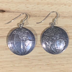 May include: A pair of silver-toned drop earrings featuring a circular pendant design. Each pendant is embossed with a detailed map of the world, including lines of latitude and longitude. The earrings are attached to standard silver-toned metal hooks.
