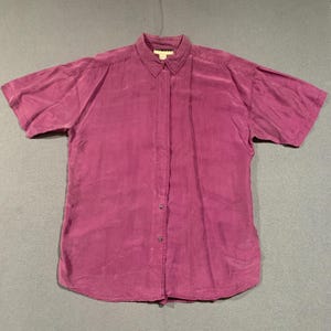 May include: A short-sleeved, button-down shirt in a deep magenta hue. The shirt features a classic collar and a vertical seam down the front. The fabric appears to have a subtle sheen, suggesting a silk or similar material.