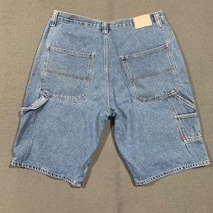 May include: Light blue denim shorts featuring a classic five-pocket design with two large back pockets, a utility loop on the left side, and a small side pocket on the right leg. A plain beige leather-like patch is attached to the back waistband.