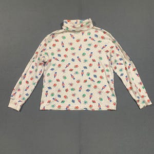 May include: White turtleneck long-sleeve shirt with a repeating pattern of gift boxes, flowers, and bottles in red, green, and blue. The shirt has a fitted silhouette and is made of a soft, lightweight fabric.