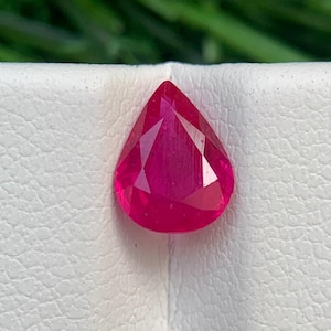 Op de afbeelding: Een gefacetteerde, peervormige robijn edelsteen. De edelsteen heeft een levendige, dieprode kleur en ligt op een wit oppervlak. De achtergrond is een wazig groen, wat op gebladerte duidt.