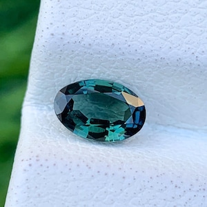 May include: A dark green oval gemstone with a faceted cut. The gemstone is set on a white background.