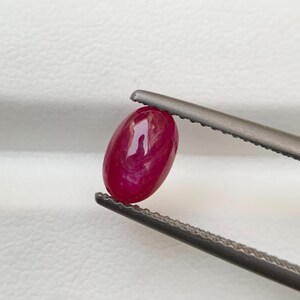 Puede incluir: Una gema de rubí ovalada, de color rojo intenso, sujeta con pinzas plateadas. El rubí muestra patrones internos. El fondo es blanco, resaltando la gema.