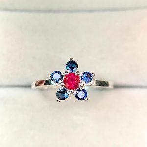 Op de afbeelding: Een zilveren ring met een bloemmotief. Het midden van de bloem is een rode edelsteen, omringd door vijf blauwe edelstenen. De ring is geplaatst tegen een zachte, neutrale achtergrond.