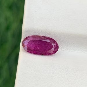 Op de afbeelding: Een gefacetteerde, ovaalvormige robijn edelsteen. De robijn heeft een diepe, verzadigde roze-rode kleur. De edelsteen ligt op een wit oppervlak met een groene achtergrond.