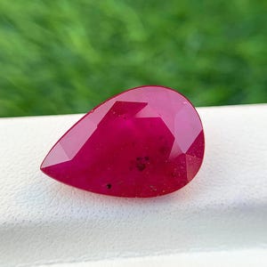 Op de afbeelding: Een diep rode peervormige edelsteen met een facetgeslepen vorm. De edelsteen heeft een lichte glans en is geplaatst tegen een witte achtergrond.