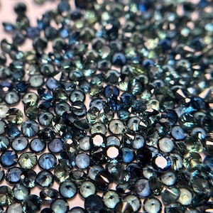 May include: A close-up view of a large quantity of loose, faceted, blue sapphires. The sapphires are various shades of blue, from light to dark, and have a slight green hue.