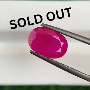 May include: An oval-cut, vibrant pinkish-red gemstone held by a pair of metal tweezers. The gemstone features multiple facets that reflect light. The text "SOLD OUT" is printed in bold black capital letters across the upper portion of the image.