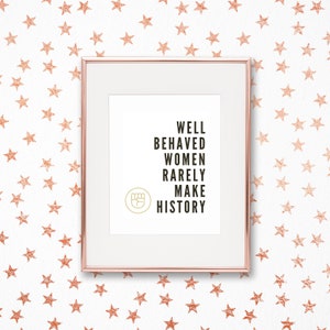 May include: A framed print with a rose gold frame and a white background. The print features the text "Well Behaved Women Rarely Make History" in black text with a raised fist icon in gold.