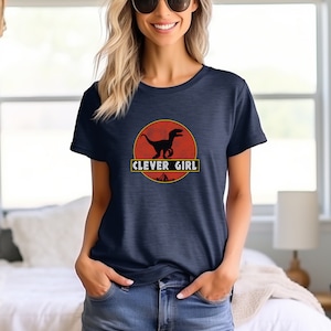 Può includere: Una maglietta blu navy con una grafica di un dinosauro in un cerchio rosso con il testo "CLEVER GIRL" sotto.