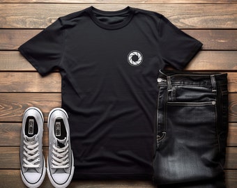 Może przedstawiać: Czarny t-shirt z białym nadrukiem obiektywu aparatu na lewej piersi. Koszulka jest zestawiona z ciemnymi jeansami i szarymi butami sportowymi.