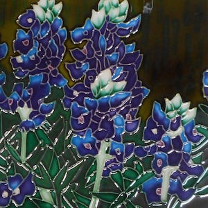 May include: A square ceramic tile featuring a detailed illustration of bluebonnet flowers. The flowers are depicted in shades of blue and purple with green leaves and white accents. The tile has a glossy finish and a dark background.