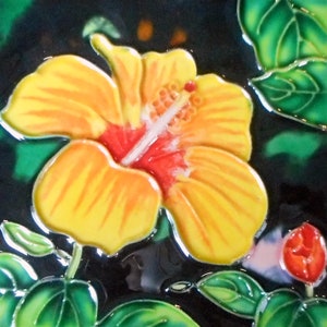 May include: A yellow hibiscus flower with a red center, painted on a ceramic tile. The flower is surrounded by green leaves.