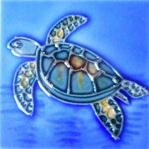 May include: A ceramic tile featuring a sea turtle design. The turtle has a blue shell with brown and yellow accents, and is set against a blue background. The tile is square and has a glossy finish.